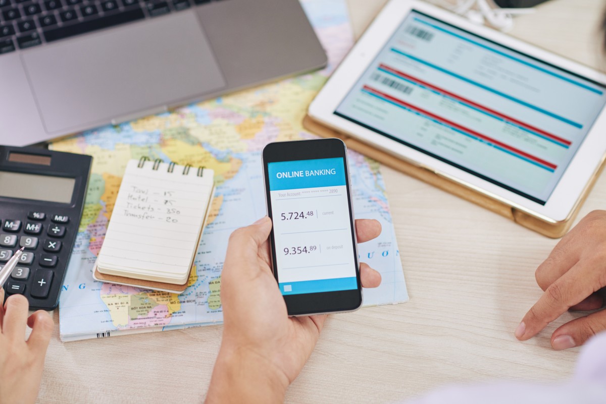 A person is holding a smartphone displaying an online banking app with account balances. The phone shows a current balance of 5,724.48 and a deposit of 9,354.89. Nearby, there's a tablet with information on the screen, a laptop, a calculator, and a notepad with travel expenses listed, including taxi, hotel, tickets, and transfer costs. The background includes a map, indicating a possible travel planning session.
