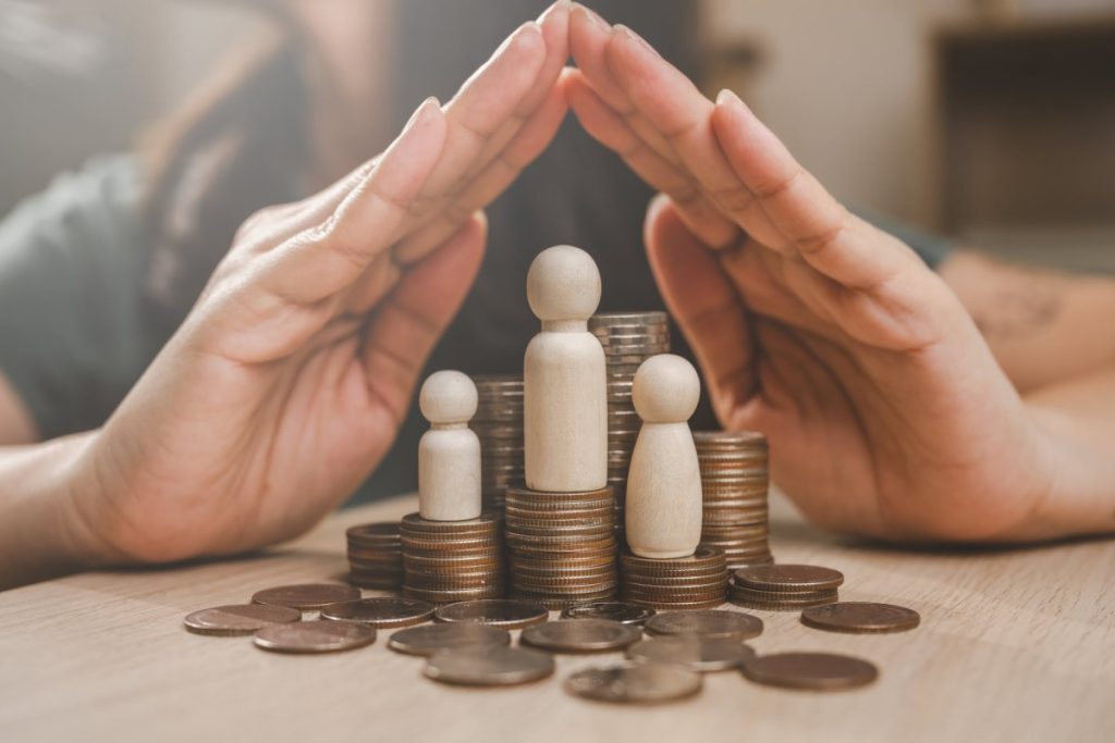 Hands protecting wooden figures on coins pile.