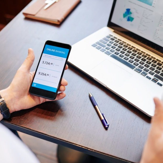A person sits at a wooden table holding a smartphone displaying an online banking app, showing account balances. An open laptop with graphs and a notebook are on the table. A pen and a takeaway coffee cup are nearby.