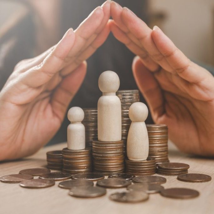 Hands protecting wooden figures on coins pile.