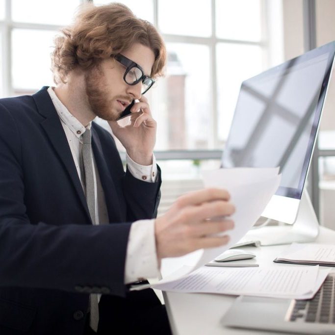 A man in a suit and tie, wearing glasses, is talking on a mobile phone while holding a sheet of paper in his other hand. He is seated at a desk in front of a computer monitor and a laptop, with various documents spread out on the desk. The office setting features large windows allowing natural light to illuminate the room.